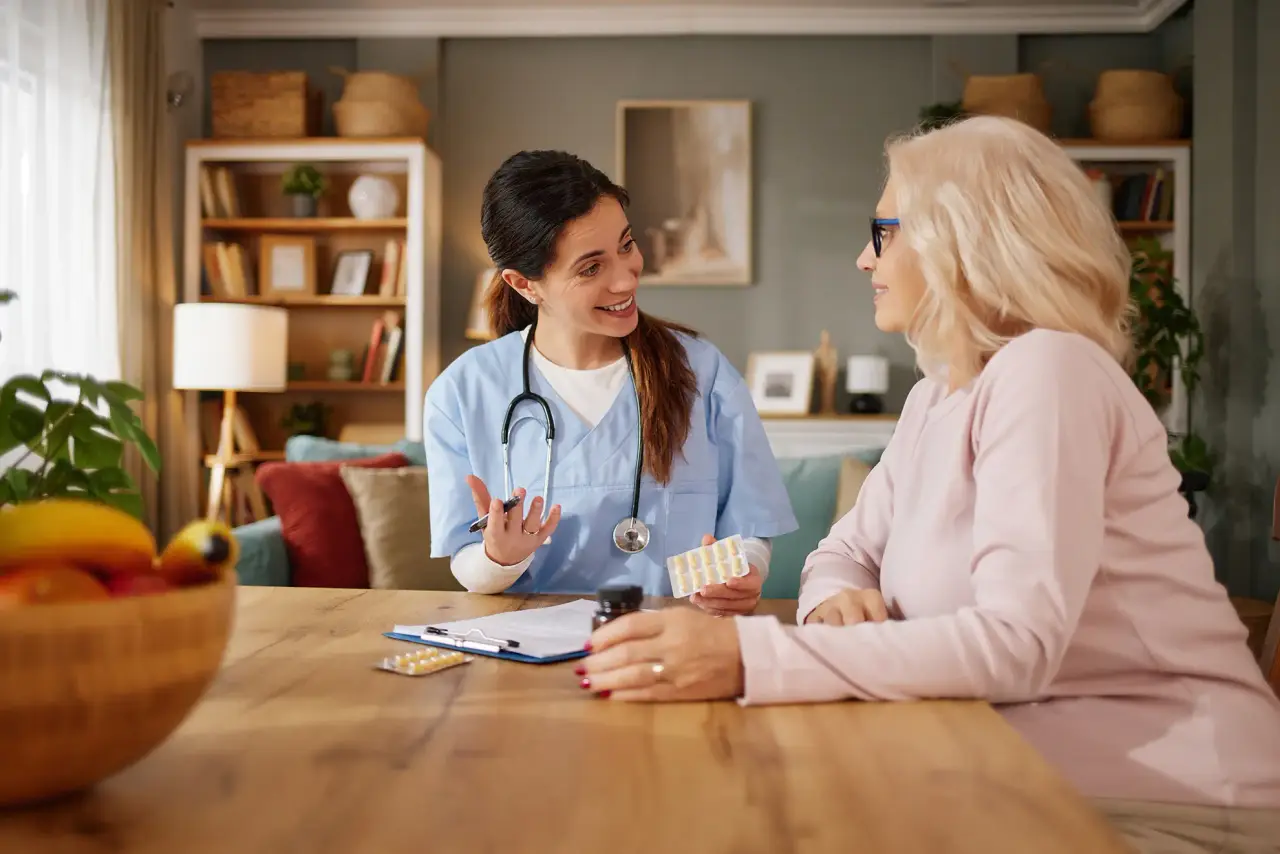 A nurse with a stethoscope talks with an older woman at a table in a bright living room.