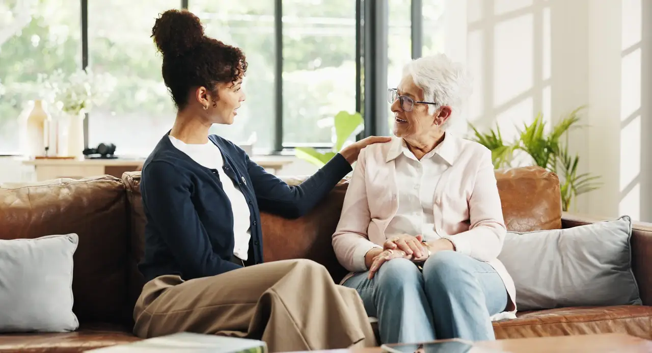 A caregiver rests a reassuring hand on the shoulder of a smiling older woman as they talk on a sofa.