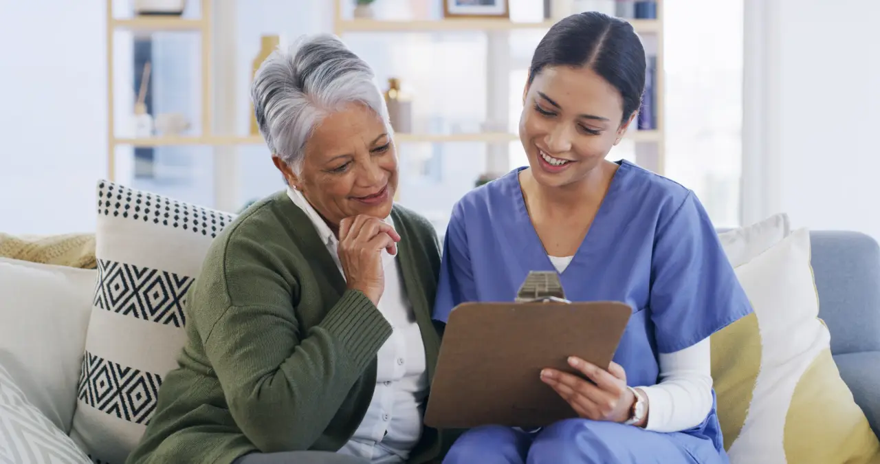 A nurse in scrubs shares a clipboard with a smiling older woman as they review a care plan together.
