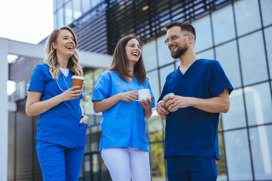 Three smiling caregivers in blue scrubs chat over coffee outside a modern building.