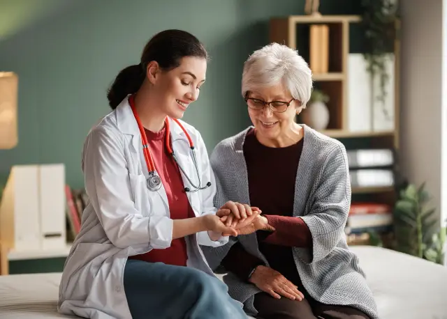A clinician in a white coat holds the hands of a smiling older woman.