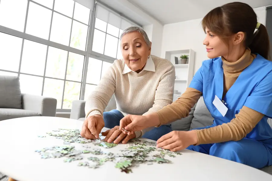 A caregiver in blue scrubs and an older woman work on a jigsaw puzzle together at a table.