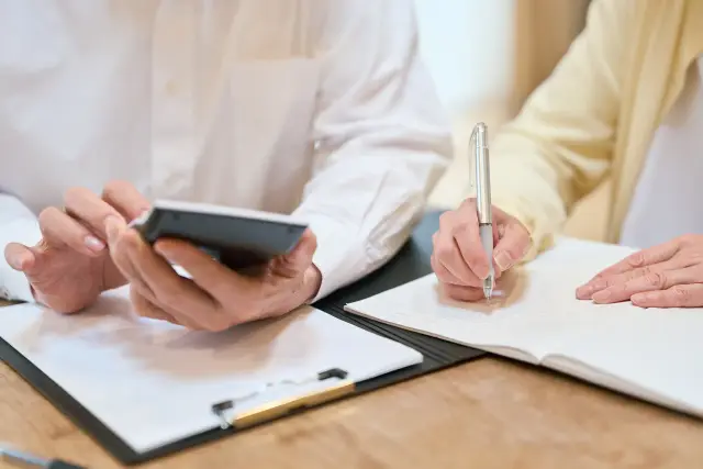 Two people review paperwork at a table with a calculator and notebook.