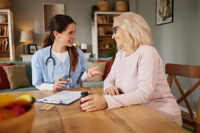 A nurse with a stethoscope talks with an older woman at a table in her living room.