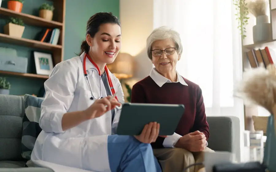 A clinician in a white coat shows a tablet to a smiling older woman in her living room.
