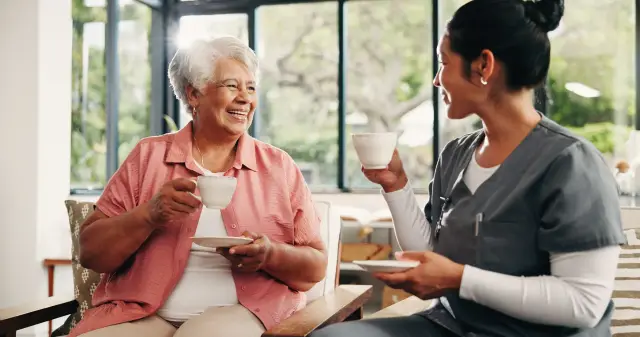A smiling older woman in a coral shirt shares a cup of tea with her caregiver.
