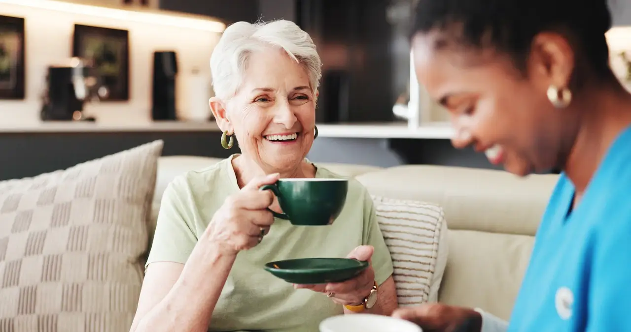 A smiling older woman enjoys a cup of coffee on the sofa beside her caregiver.