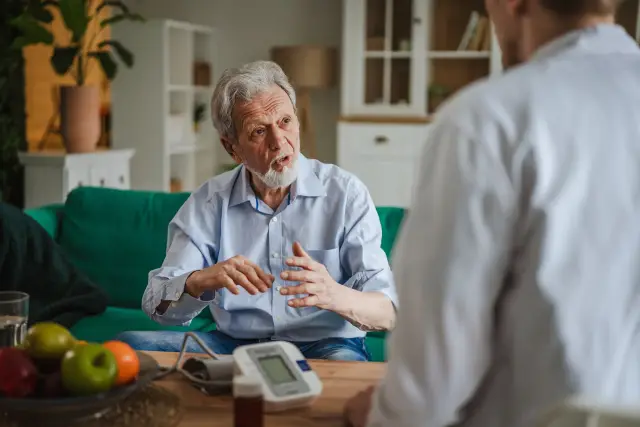 An older man talks with a nurse at home, with a blood pressure monitor on the table.
