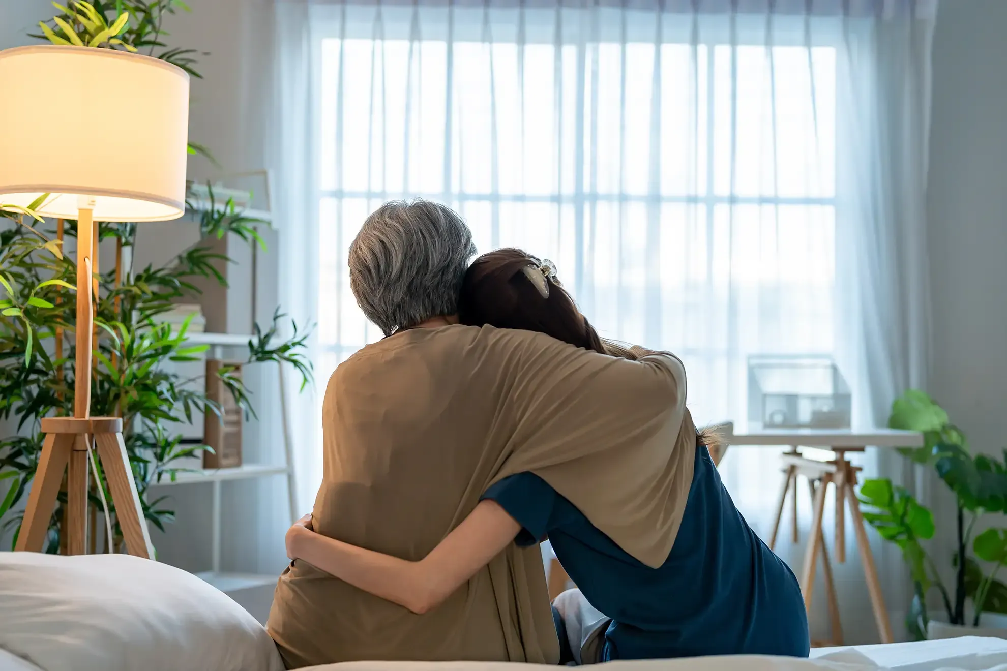 A daughter hugs her older mother as they sit together on a bed looking toward a bright window.