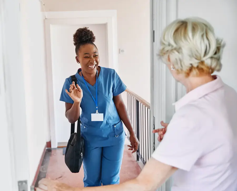 A smiling caregiver in blue scrubs is welcomed at the front door by an older woman.