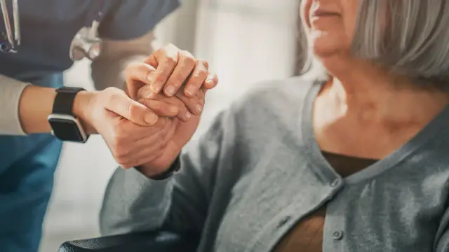 A caregiver gently holds the hand of an older woman in a grey cardigan.