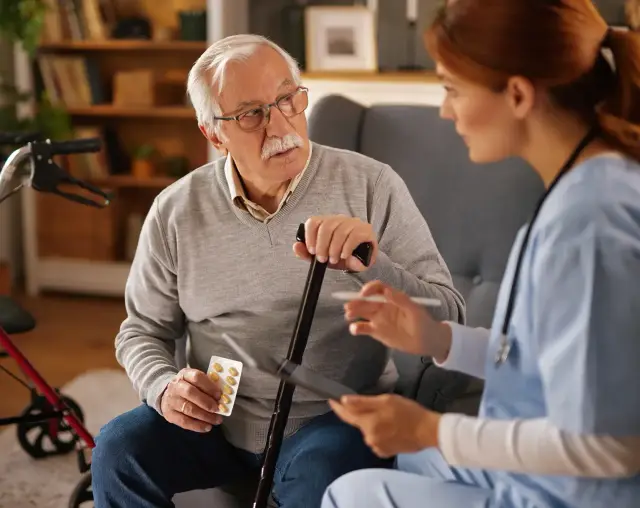 An older man holding a cane and a blister pack of pills listens to a clinician seated beside him.
