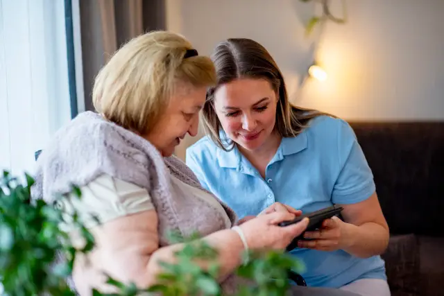 A caregiver and an older woman sit close together on a sofa, smiling as they look at a phone.