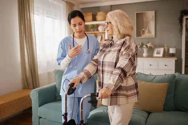 A nurse in scrubs guides a woman using a walker through her living room.