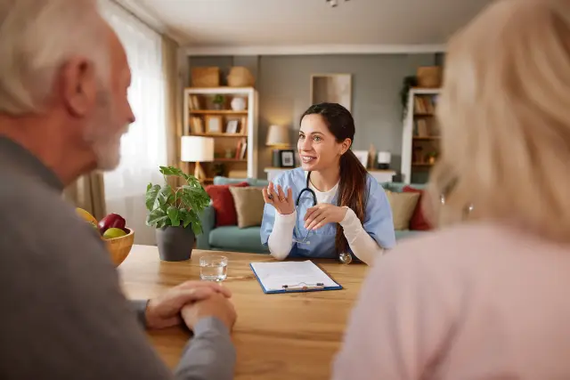 A nurse talks with an older couple across a table in their living room.