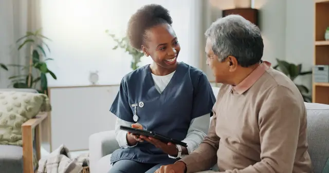 A smiling nurse holding a tablet talks with an older man on a sofa.