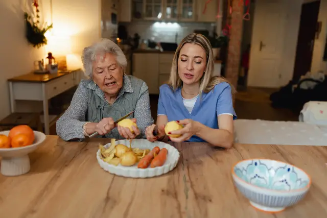 A caregiver and an elderly woman peel vegetables together at a wooden kitchen table.