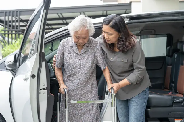 A woman helps an elderly woman with a walker step out of a vehicle.
