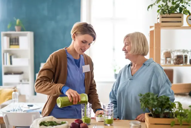 A caregiver pours a green smoothie in the kitchen while an older woman looks on, smiling.