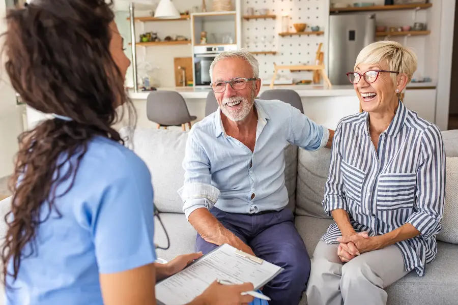 A nurse with a clipboard shares a laugh with an older couple seated on their sofa.