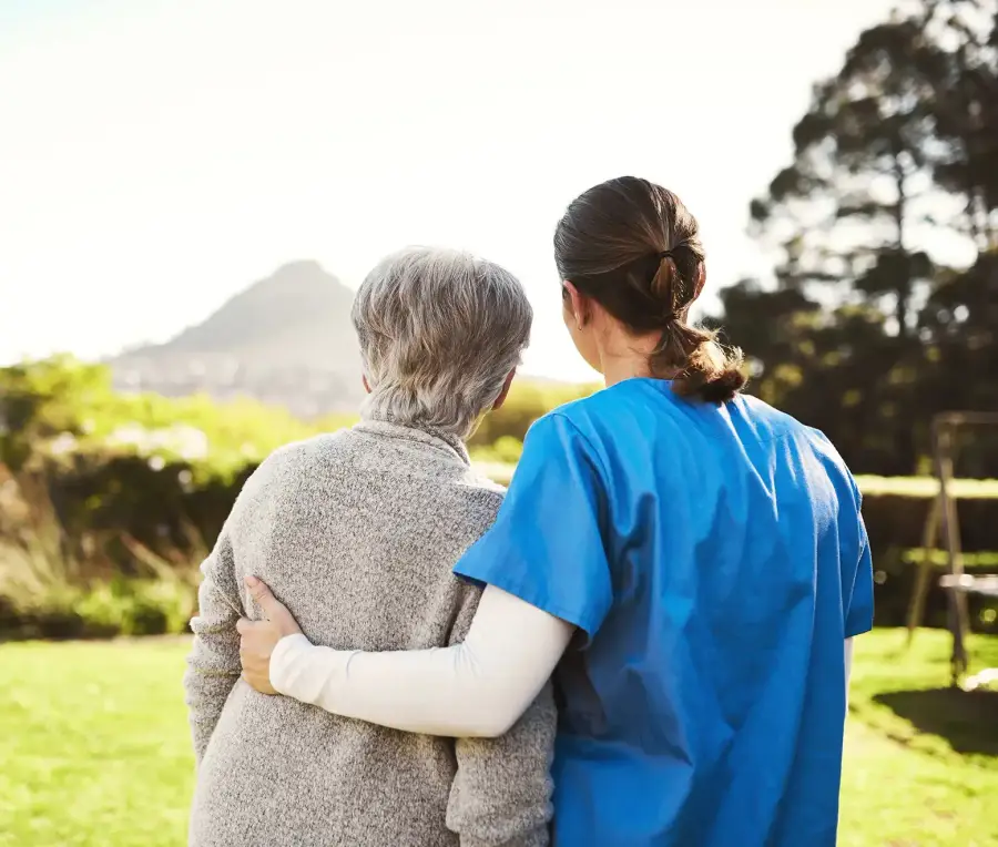 A caregiver in blue scrubs with her arm around an older woman, seen from behind as they look outdoors.