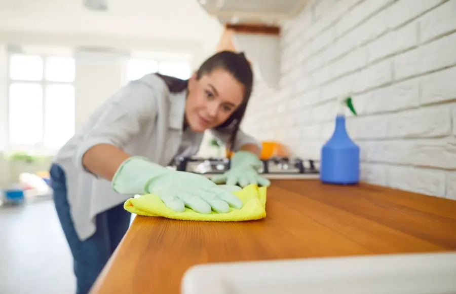 A caregiver wearing gloves wipes down a wooden kitchen counter with a yellow cloth.