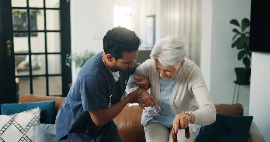 A caregiver supports an older woman by the arm as she rises from a sofa with her cane.