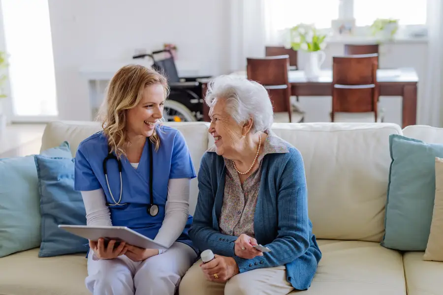 A nurse with a stethoscope and tablet laughs with an elderly woman on a sofa at home.