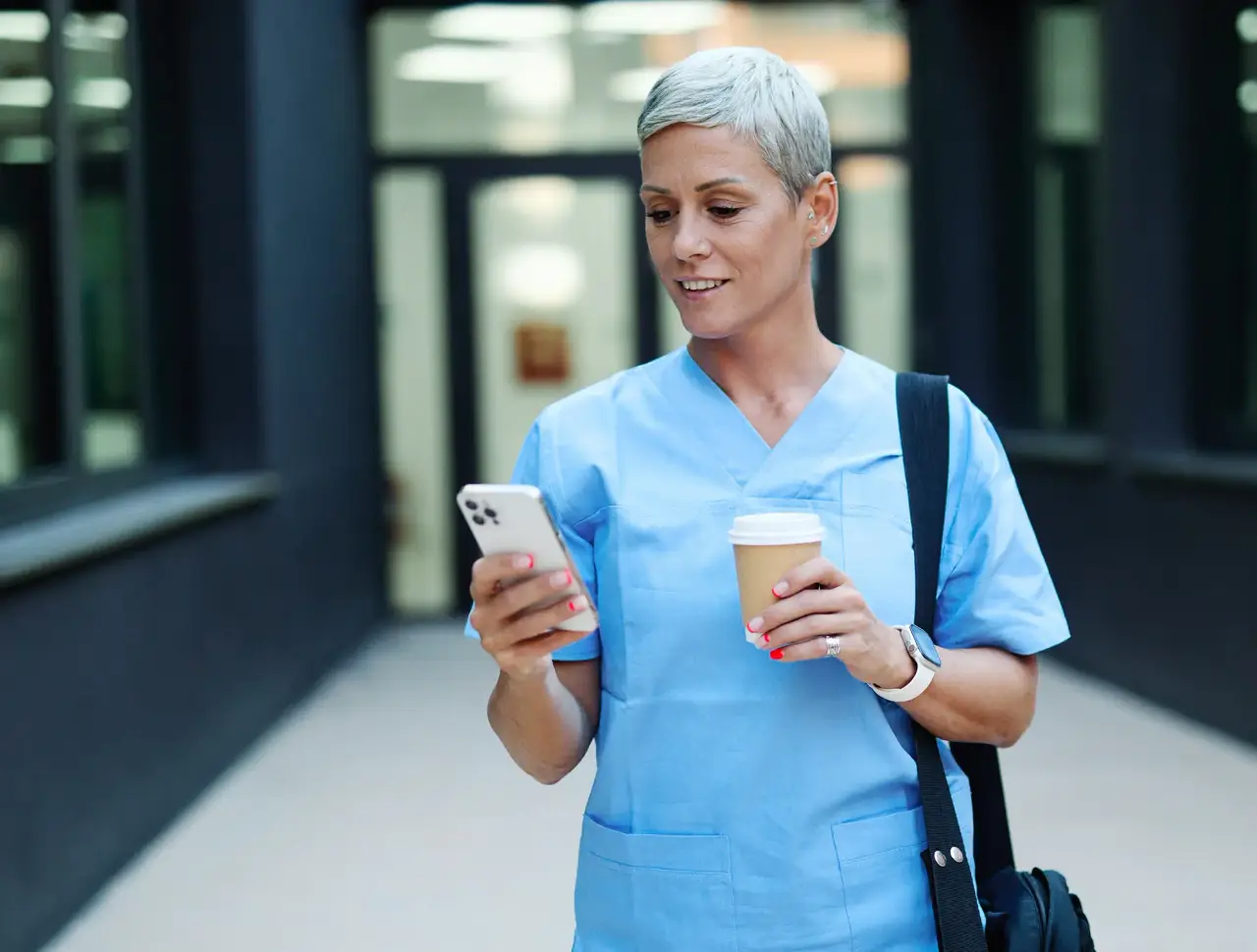 A nurse in blue scrubs checks her phone while holding a coffee outside a building.