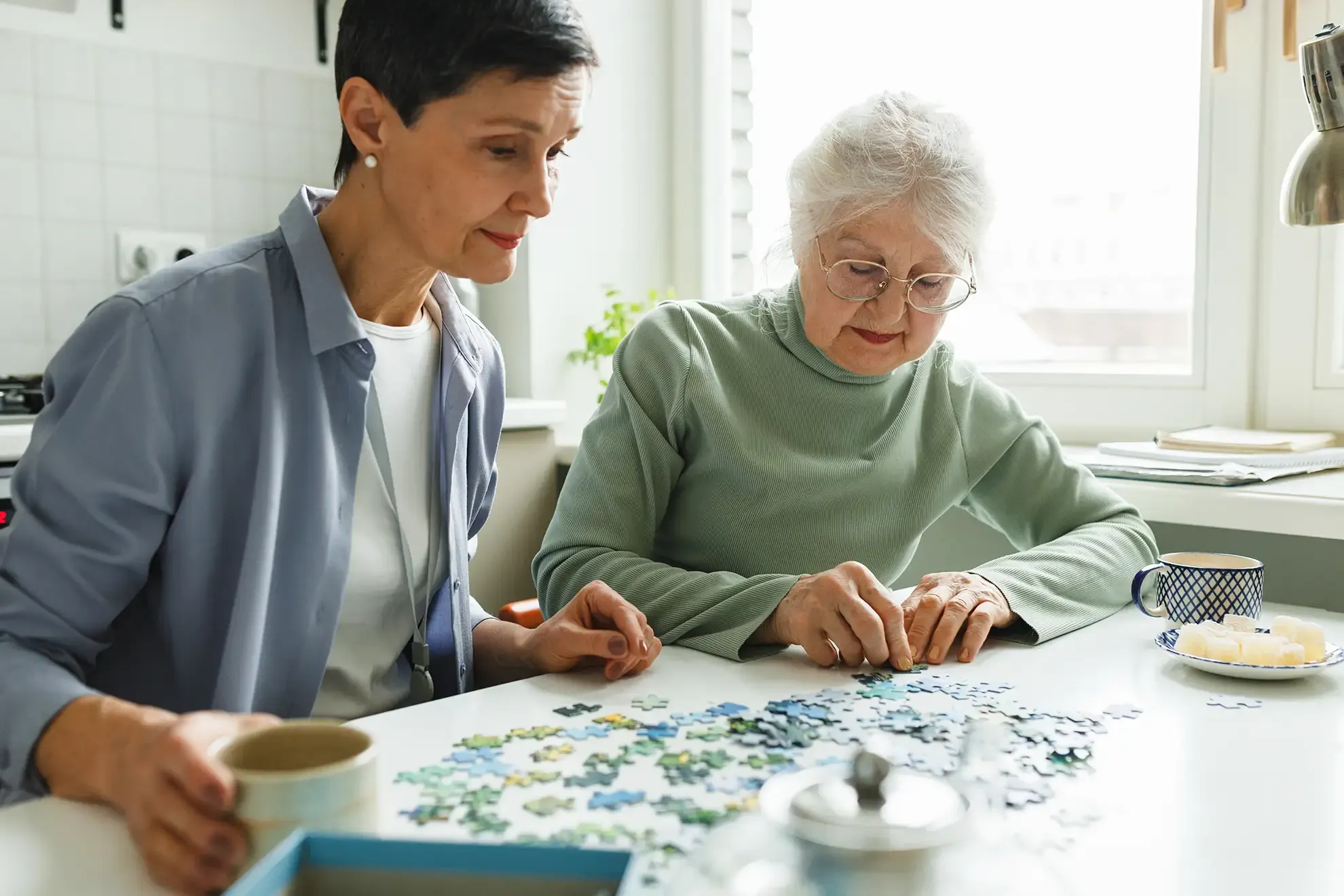 A woman and her elderly mother work on a jigsaw puzzle together at a bright kitchen table.