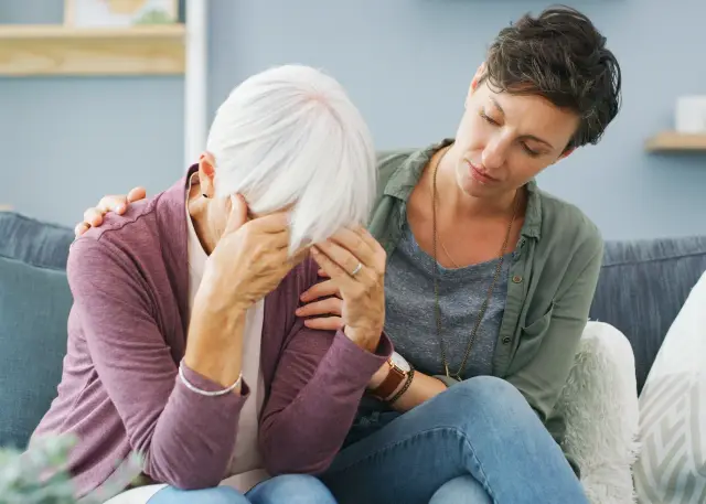 A woman comforts her older mother, who holds her head in her hands, on a sofa.