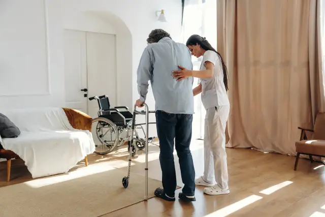A caregiver steadies a man as he walks with a cane through a bright living room, a wheelchair nearby.