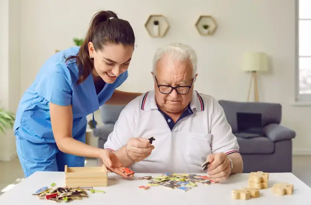 A caregiver assists an elderly man with colorful puzzle pieces on a table, both engaged in a thoughtful activity in a bright room.