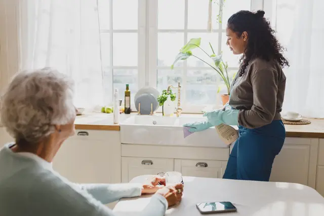 A woman with curly hair cleans a kitchen sink while an older woman enjoys a drink at the table.