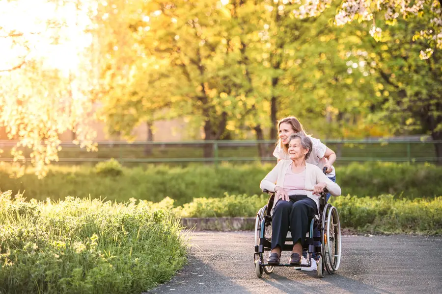 A young woman pushes an elderly woman in a wheelchair along a sunlit path surrounded by lush greenery.