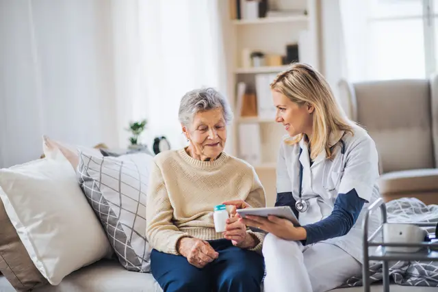 A caregiver engages warmly with an elderly woman, discussing something on a tablet while seated in a cozy living room.