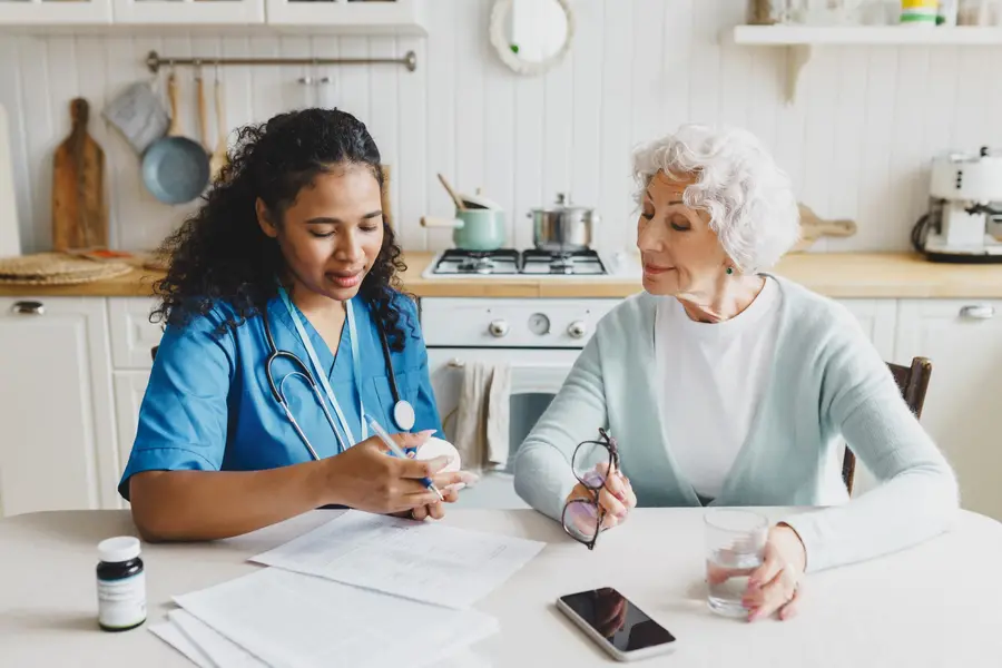 A healthcare professional discusses medication with an elderly patient at a kitchen table, emphasizing care and communication.